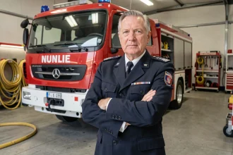 Pompier senior en uniforme de cérémonie avec insignes de grade devant un camion de pompiers rouge en caserne urbaine